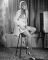 Wendy Richard, wearing a light-colored mini-dress and long wig, sitting on a barstool